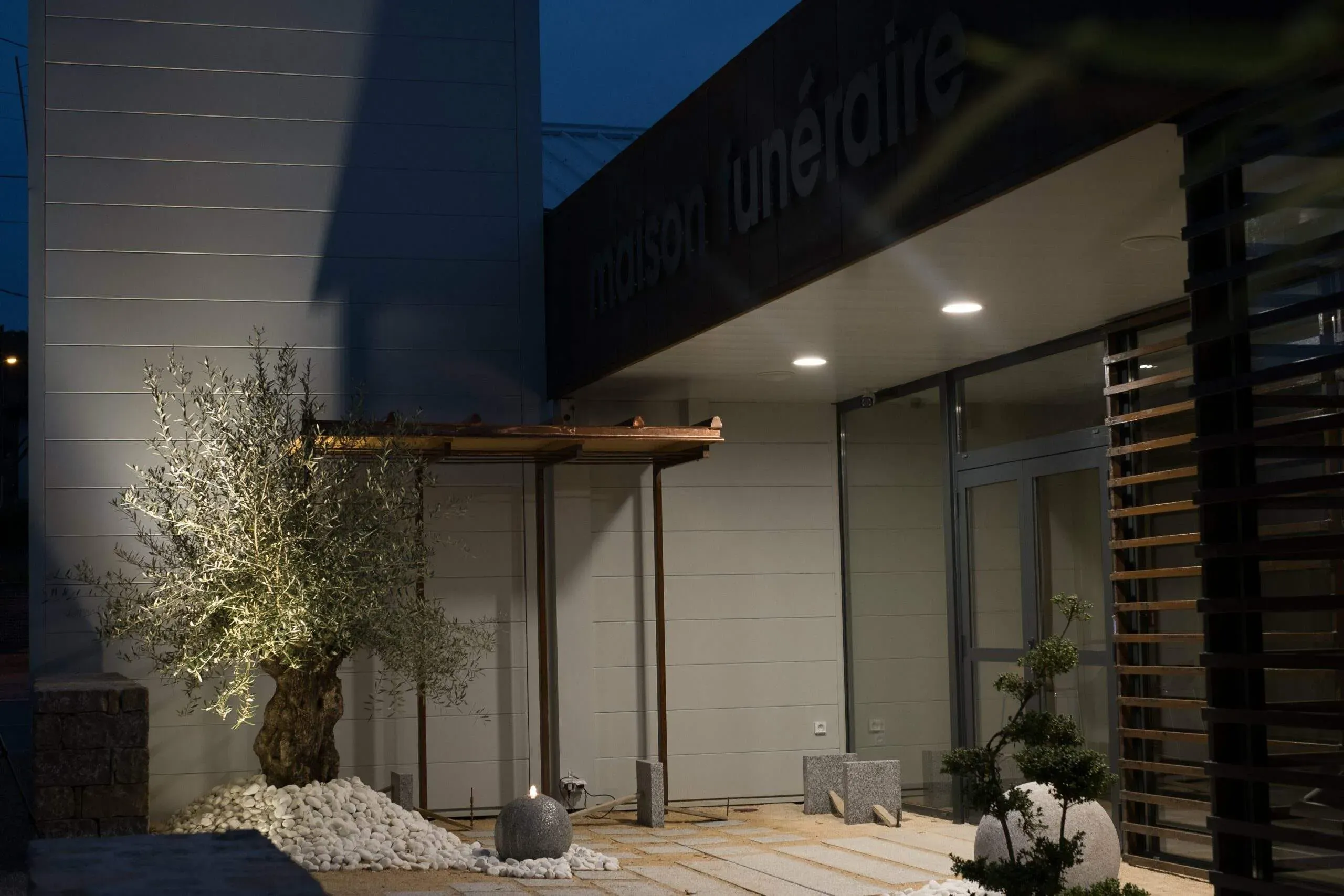 Façade moderne de la Maison Funéraire avec olivier éclairé et décorations zen.